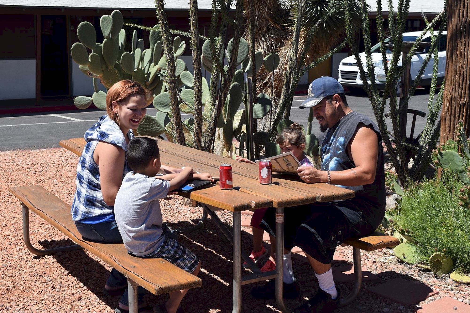A family is seated at a picnic table outside, surrounded by cacti, enjoying drinks and snacks on a sunny day.
